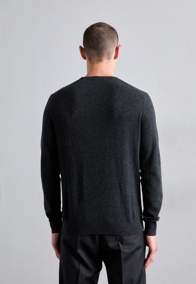 Man with short hair facing away wearing a long-sleeve dark gray sweater and black pants against a plain light gray background.