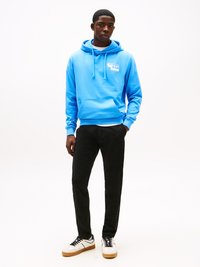 Lichtblauwe hoodie met een voorzak, met een wit logo. Gecombineerd met zwarte broek en witte sneakers met een beige zool en zwarte veters.