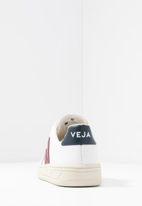 Zapatilla de piel blanca con una pestaña en el talón azul etiquetada "VEJA", acentos laterales rojos y una suela texturizada color crema. La vista trasera muestra un diseño minimalista.