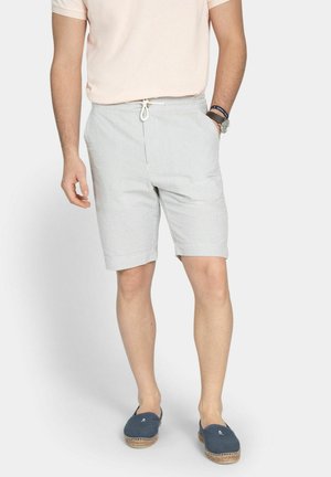 Mand iført lyserød skjorte, lysegrå stribede shorts med snøre, marineblå espadriller, armbåndsur og armbånd, stående med afslappede hænder.