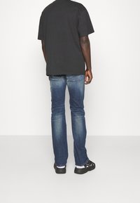 Person som bär urtvättade blå jeans, svart t-shirt, vita strumpor och svarta sandaler, stående framför en enkel vit bakgrund och vänder ryggen mot kameran.