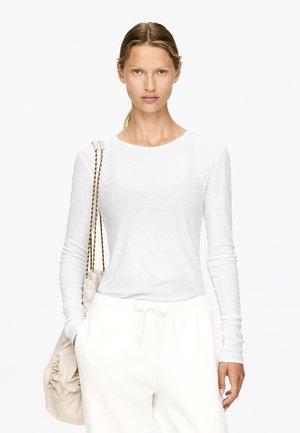 Langärmliges weißes Oberteil aus leichtem Stoff, runder Ausschnitt, kombiniert mit weißen Jogginghosen mit Kordelzug und einer cremfarbenen Tasche mit geflochtenen Trägern.