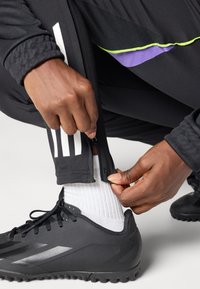 Svarte treningsbukser med hvite striper, med glidelåser ved anklene, justeres av en hånd, sammen med svarte fotballsko og hvite sokker.