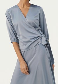Top avvolgente in satin blu chiaro con maniche tre-quarti, annodato in vita. Tessuto liscio, scollatura a V e dettagli sulle maniche arricciate.