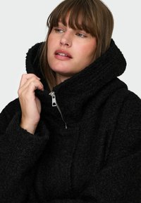 Črna flis bunda z visokim ovratnikom, polovičnim zadrgo in rebrastimi detajli. Mehek tekstura, sproščen kroj in brez dodatnih vzorcev ali okrasitev.