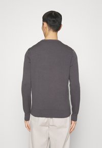 Mężczyzna stojący tyłem, ubrany w ciemnoszary sweter z długim rękawem i jasnobeżowe spodnie na jednolitym białym tle.