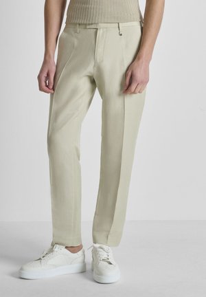 Persona vestida con pantalones beige a medida, zapatillas blancas y una camiseta sin mangas beige de punto acanalado, de pie contra un fondo claro y sencillo.