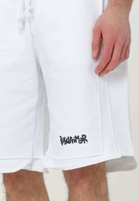 Pantaloni corti bianchi in tessuto morbido, con orlo grezzo, tasche laterali e logo "DISCLAIMER" in nero sul lato sinistro in basso.