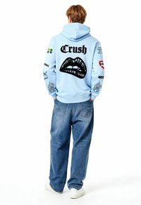 Felpa con cappuccio azzurro chiaro con una grande grafica nera di labbra e la scritta "Crush" sul retro; presenta varie grafiche sulle maniche; indossata con jeans larghi.