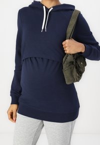 Marineblauwe hoodie met een cropped ontwerp, elastische onderrand en trekkoord. Gekoabd met lichtgrijze sweatpants en een textuurachtige olijfgroene tas.