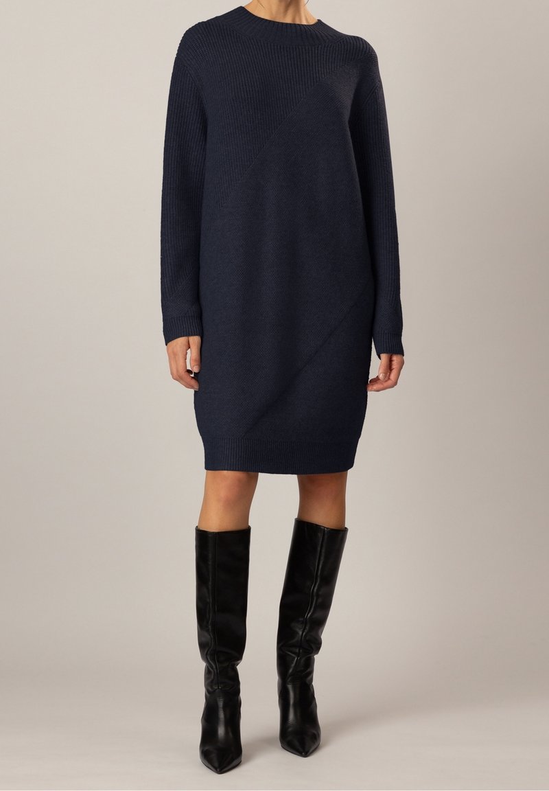 Femme portant une robe en maille texturée bleu marine foncé longueur genou avec des manches longues, associée à des bottes en cuir noires montant jusqu'aux genoux.