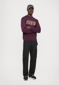 Felpa bordeaux con "VERSACE JEAN COUTURE" in lettere dorate, abbinata a pantaloni cargo neri e sneakers nere. Un cappellino grigio completa il look.