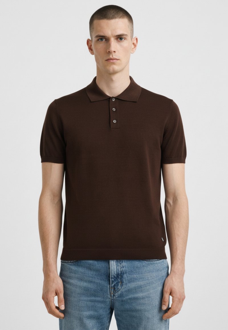 Młody mężczyzna ubrany w ciemnobrązową koszulkę polo z krótkim rękawem i jasnoniebieskie jeansy, stojący na tle jasnego, jednolitego tła.