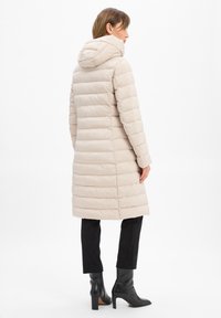 Beige Daunenjacke mit Kapuze, mit horizontalen Steppnähten, bis zur Mitte des Oberschenkels reichend. Getragen über schwarzen Hosen und Ankle Boots.