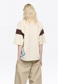 Kratka majica s kratkimi rokavi v kremni barvi, z širokimi rjavimi črtami, mehko teksturo in sproščenim krojem. Zadaj ima modro akcentacijo ob straneh.