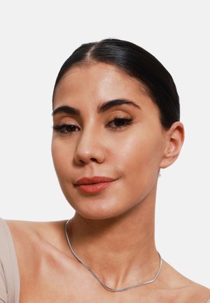 Silberne Metallkette mit einem schlanken, gebogenen Design. Die Trägerin hat glattes, dunkles Haar, minimales Make-up und einen hellen Hautton.