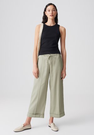 Woman wearing black sleeveless top, light green wide-leg pants, cream loafers, and a textured shoulder bag against a plain light background.