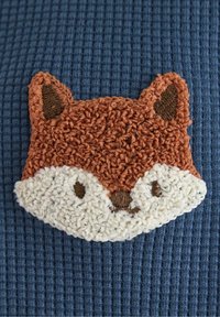 Broderad rävpatch i brunt, krämfärgad och svart på texturerad blå tyg, med distinkta öron, ögon och en ludden kant.