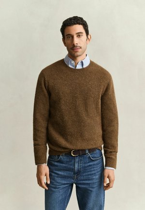 Man wearing brown textured sweater over blue collared shirt and blue jeans standing against plain light background.