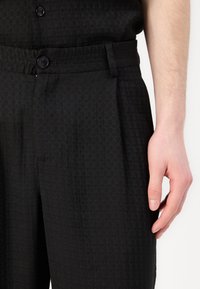 Pantalons noirs texturés avec un subtil motif circulaire, dotés d'une fermeture à bouton et de poches latérales. Le matériau semble léger et tissé.