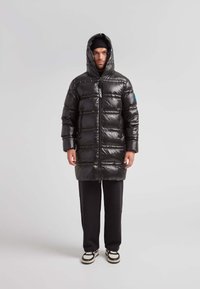 Chaqueta acolchada negra con acabado brillante, cierre de cremallera, capucha de gran tamaño y diseño acolchado horizontal, combinada con pantalones negros y zapatillas.