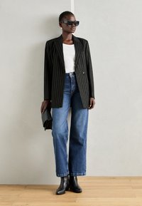 Kvinne iført en svart pinstripe blazer, hvit skjorte, blå vide jeans, svarte støvletter, store solbriller, gulløredobber, og holder en svart clutch.