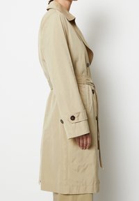 Persona che indossa un trench beige con polsini abbottonati e cintura, mostrata di lato su uno sfondo bianco uniforme.
