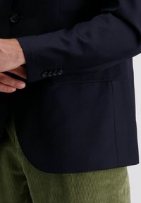 Man die de manchet van een donkerblauwe blazer aanpast, gedragen over groene ribbroeken, tegen een neutrale achtergrond.