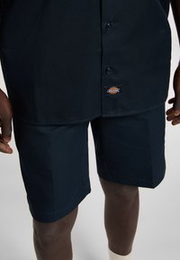Camicia blu navy realizzata in un tessuto liscio con un design a bottoni, abbinata a pantaloni corti blu navy caratterizzati da un taglio semplice e una texture sottile.