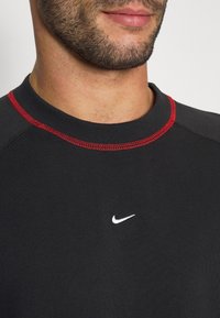Muž oblečený v černém tričku s červeným prošíváním u výstřihu a malým bílým logem Nike na hrudi, viditelná čelisti a oblast krku.