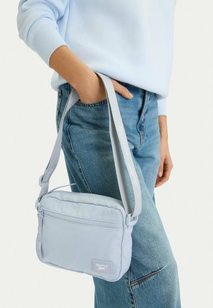 Persona que lleva una sudadera azul claro y vaqueros sostiene una bolsa cruzada gris claro de Reebok con correa ajustable a un lado.