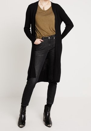 Personne portant un long cardigan noir, un t-shirt vert olive à col en V, un jean noir taille haute et des bottines noires à bouts métallisés.