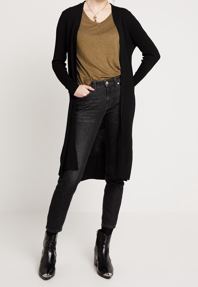 Personne portant un long cardigan noir, un t-shirt vert olive à col en V, un jean noir taille haute et des bottines noires à bouts métallisés.