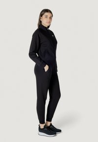 Tuta sportiva nera con colletto zip, design aderente, tasche laterali e polsini a coste; abbinata a sneaker nere con suole bianche.