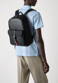 Sac à dos en cuir noir avec un rabat et une fermeture à boucle. Il dispose de bretelles rembourrées et d'un accent subtil rouge, blanc et bleu.