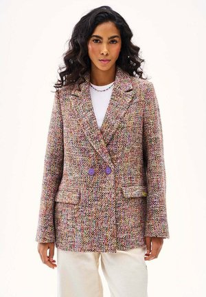 Kobieta o długich, kręconych włosach nosi wielokolorową marynarkę z tweedu z fioletowymi guzikami, pod którą ma biały top, oraz kremowe spodnie, stoi na tle jednolitego tła.