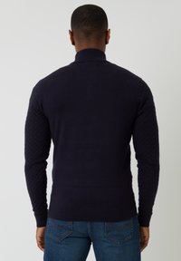 Granatowy sweter z wysokim kołnierzem, teksturowanymi, pikowanymi rękawami i dopasowanym krojem. Noszony z dżinsowymi spodniami.