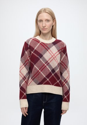 Frau trägt einen langärmeligen Pullover mit roten, beigen und violetten Rautenmuster und dunkle Hosen vor einem weißen Hintergrund.