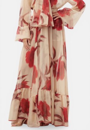 Abito maxi fluido beige con grande stampa floreale rossa, maniche lunghe larghe, cintura in vita e orlo arricciato, indossato con tacchi con cinturino trasparente.