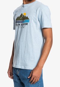 Hombre con una camiseta Quiksilver de color azul claro con un gráfico de montaña, sol y ola, combinada con jeans azules, frente a un fondo blanco.