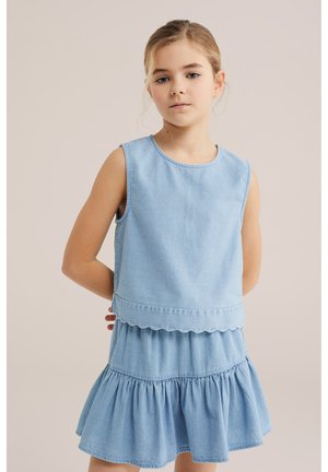 Mladé dievča s blond vlasmi zviazanými vzadu, oblečené v svetlomodrom rifľovom topu bez rukávov a zodpovedajúcej vrstvenej sukni, stojace s rukami za chrbtom.