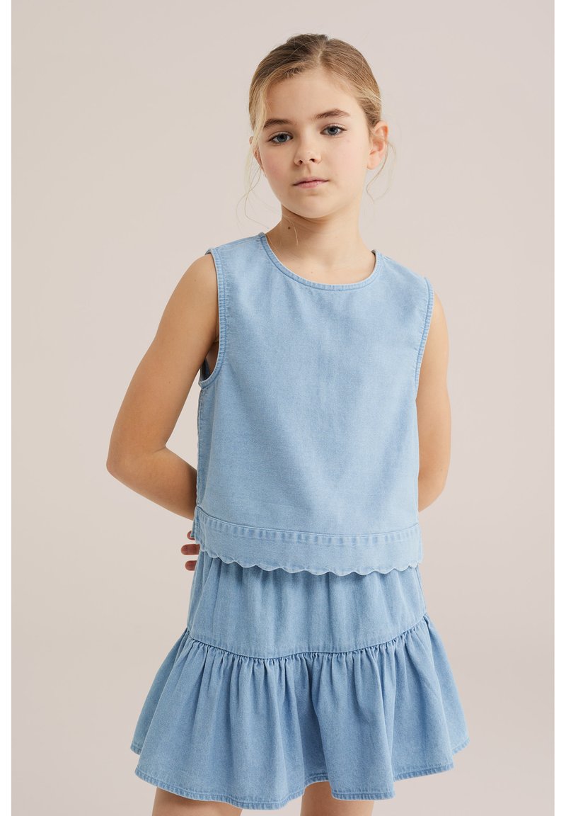 Jeune fille aux cheveux blonds attachés, portant un haut en jean bleu clair sans manches et une jupe à volants assortie, debout les mains dans le dos.