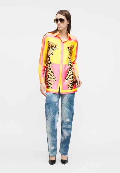 Γυναίκα που φορά υπερμεγέθη κίτρινο και ρο ζ μακρύ πουκάμισο με σχέδιο λεοπαρ, ξεβαμμένα μπλε τζιν, μαύρες γόβες και αεροπορικά γυαλιά ηλίου, όρθια.