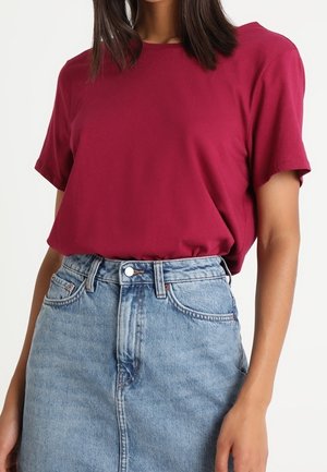Mujer con una camiseta burdeos de manga corta metida por dentro y una falda vaquera azul de cintura alta sobre un fondo liso.