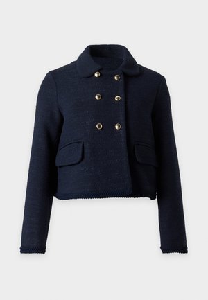 Femme portant une veste noire à double boutonnage, un jean bleu à jambes larges avec des motifs blancs, des chaussettes rouges et des chaussures plates noires, debout devant un fond blanc.