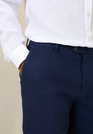 Férfi tengerészkék nadrágot és fehér, hosszú ujjú, gombolós inget visel, ballal a nadrágzsebben.