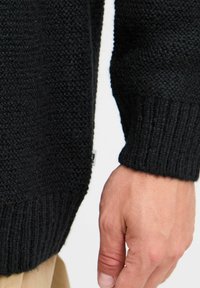 Pulover negru tricotat cu un finisaj texturat, manșete cu nervuri și detalii cu etichetă pe lateral, evidențiind un aspect dens și cald.