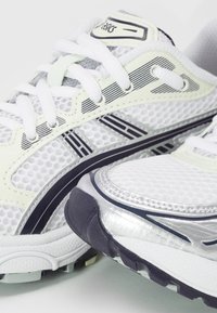 Chaussures de course Asics blanches et argentées avec des bandes noires, tissu mesh et lacets blancs, présentées en gros plan.