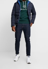 Chaqueta acolchada azul marino con acentos en azul, sudadera verde con logo, jeans de mezclilla oscuros y zapatillas blancas. Textura suave y diseño en capas.