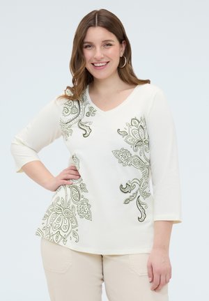 Donna sorridente con capelli castani che indossa una maglia bianca a scollo a V con motivi paisley verdi e pantaloni beige, posando con una mano sul fianco.